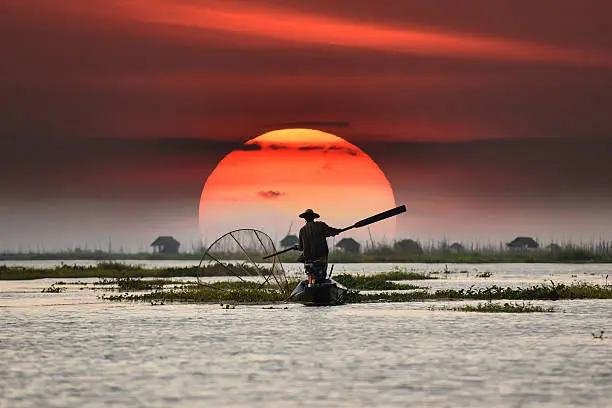 A fisherman on calm water silhouetted against a large setting sun